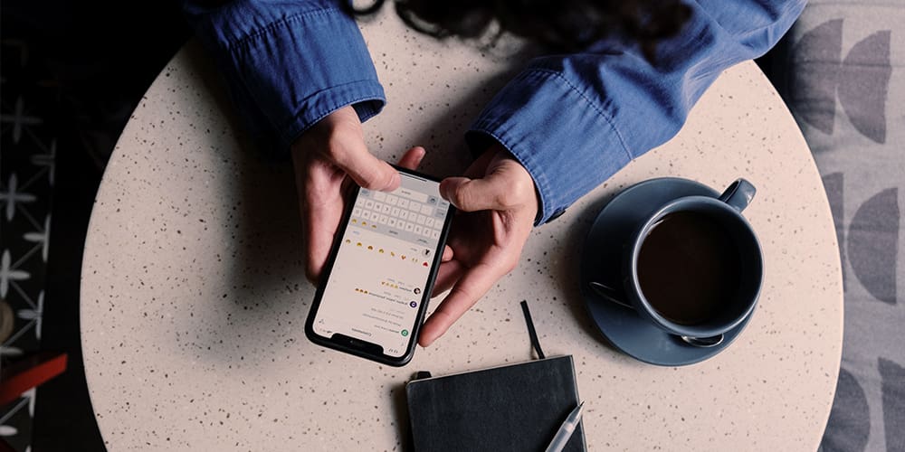Woman holding phone open to Snapchat chat window, casually dressed with journal and coffee nearby.
