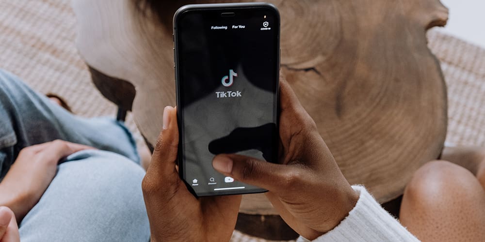 Black person's hands holding a phone loading TikTok above a wooden table.