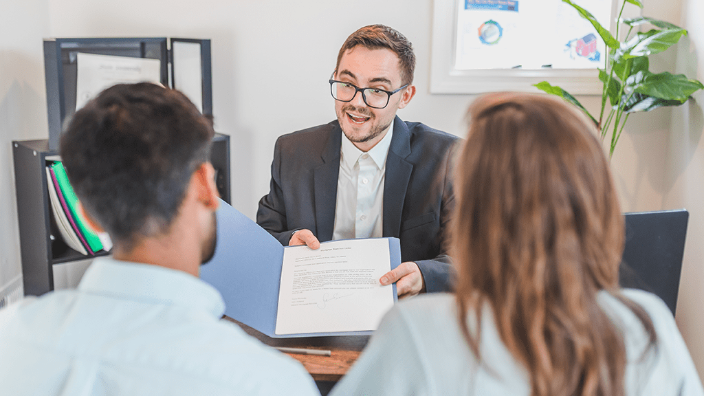 A man holds out a folder with documents in it for a man and a woman to look at while he talks in negotiation for additional duties.