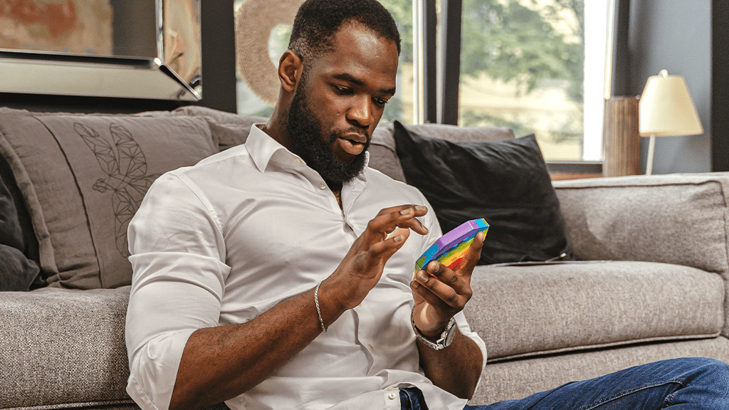 A Black man sits on the floor in front of a couch with a rainbow pop toy in his hand. He is smiling and enjoying his adult toys.