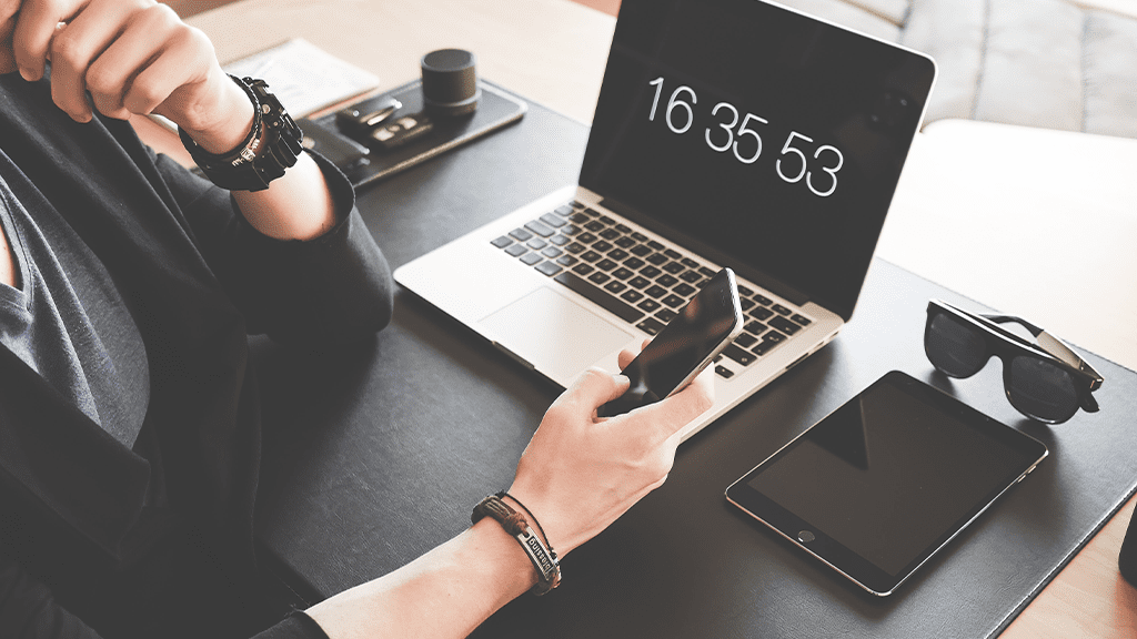 A cutoff person is holding an iPhone in their hand, with a Mac computer and an iPad behind them, showing all their Apple products.