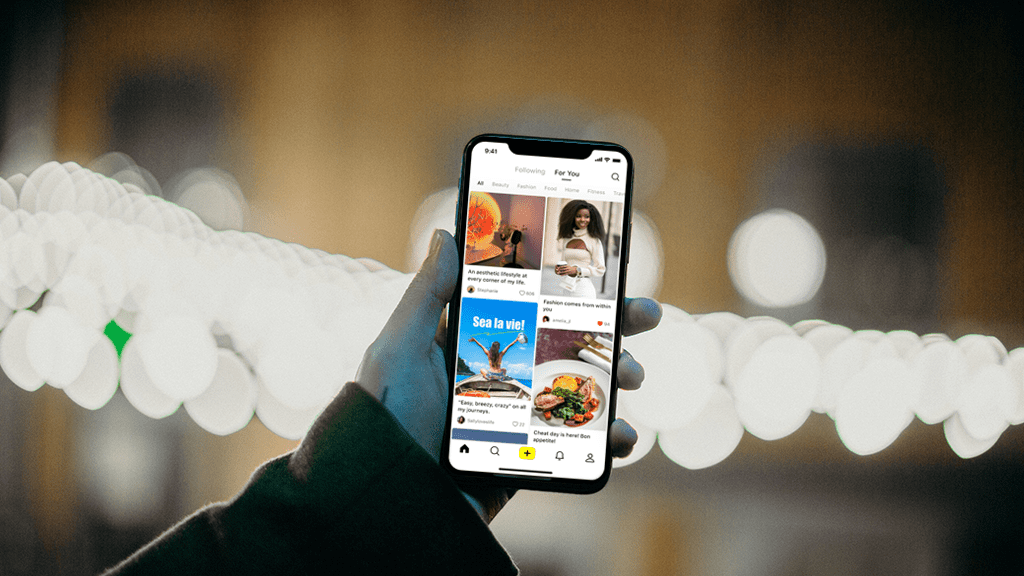 Lemon8 A hand holding a phone up against a nighttime scene with stringlights. The phone is open to the Lemon8 app from Bytedance.