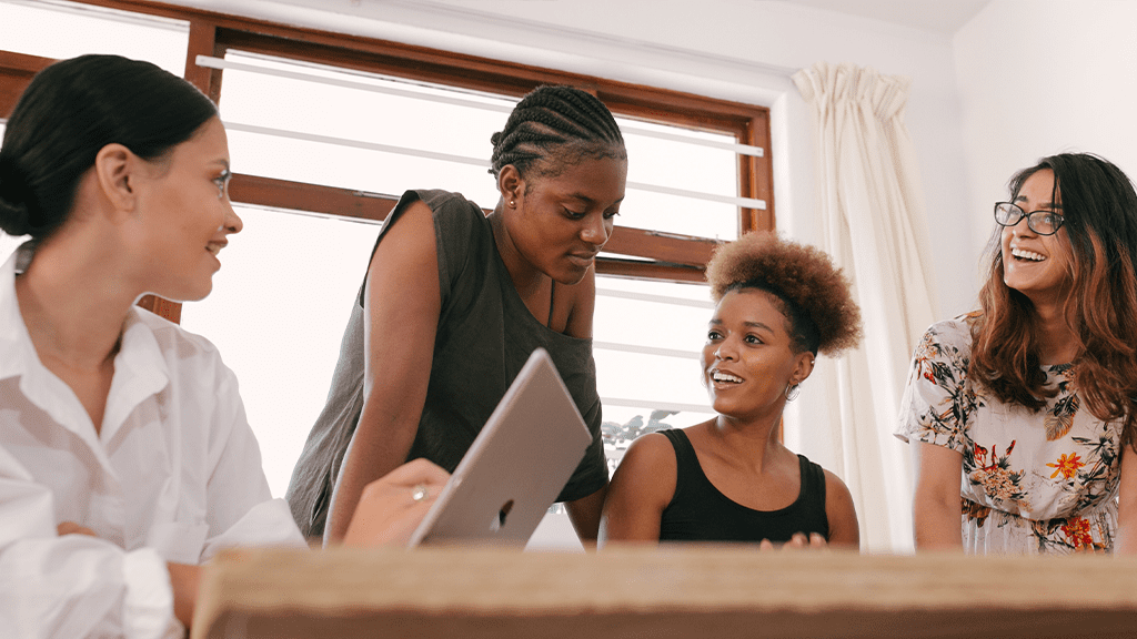 A group of women working together and standing over a table as part of the labor force, chatting animatedly.
