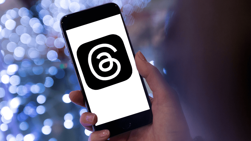A person holds up a phone with the Threads logo (a swirling at sign on a black background). A glittering blue background contrasts the phone.