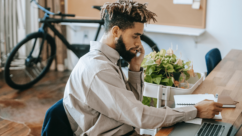 A man sits in a casual office talking on the phone and writing in a notebook.