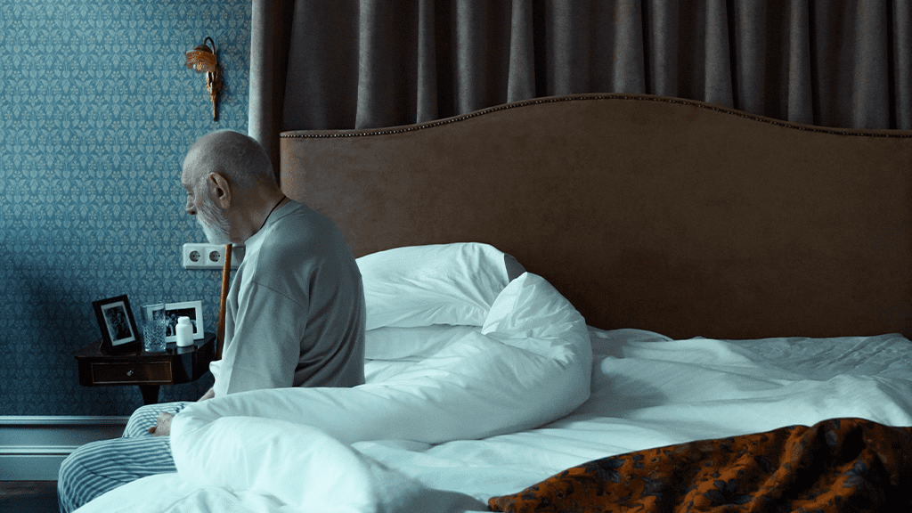 An older man sitting on the edge of a hotel bed with a heavy sad expression as he ponders an EEOC lawsuit.