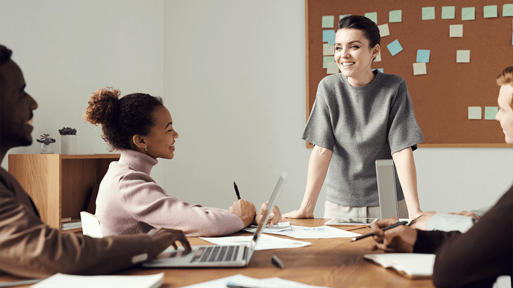 A meeting of people at an office, with a woman standing at the front of the table, welcoming ideas by leaning forward during a four day workweek.