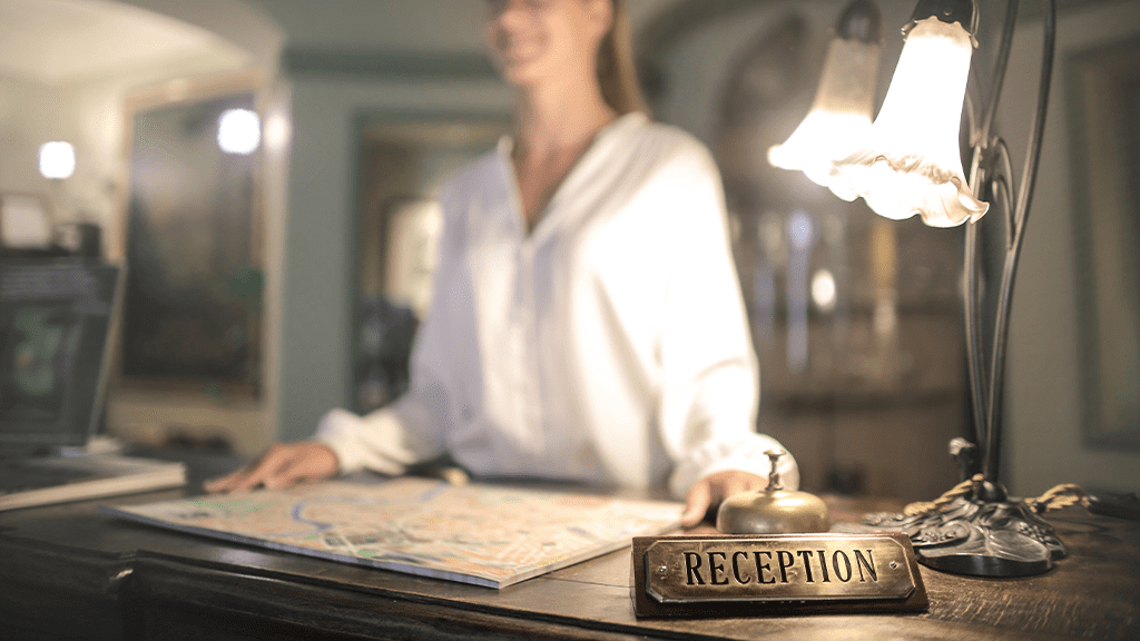 A woman standing at a hotel desk, with the focus on the foreground with the sign 'reception' illuminated by a frilled white desk lamp. The desk shows no ADA accommodation for the receptionist.