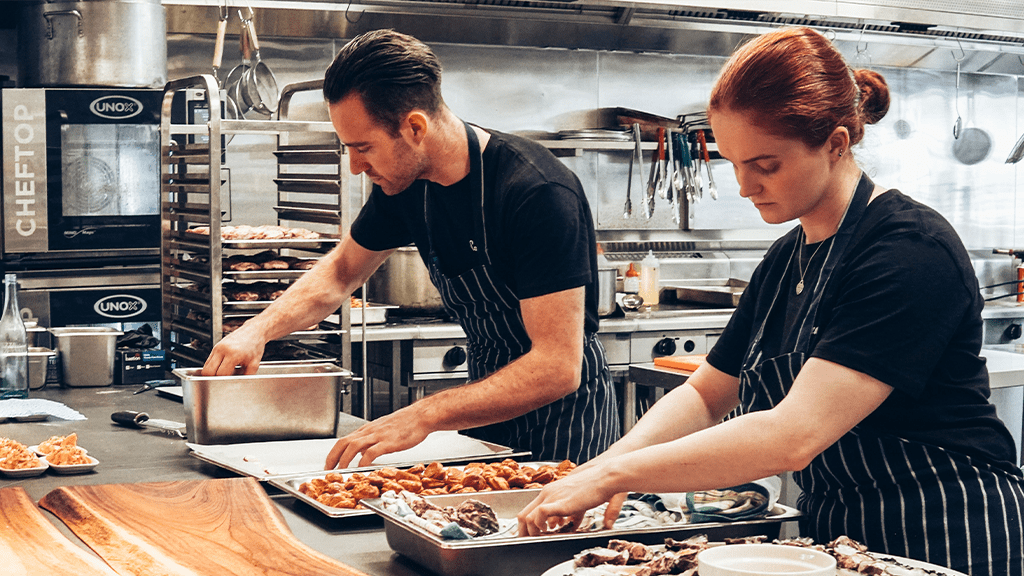 A man and a woman working in a kitchen plating food and avoiding harassment.