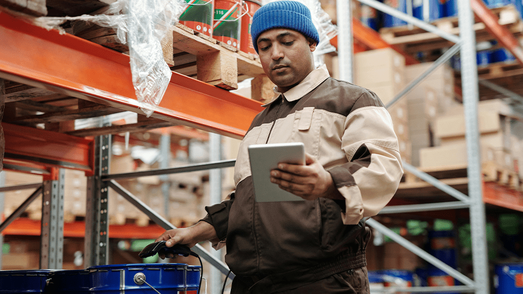 A man working in a warehouse and consulting a tablet and tool for accommodations.