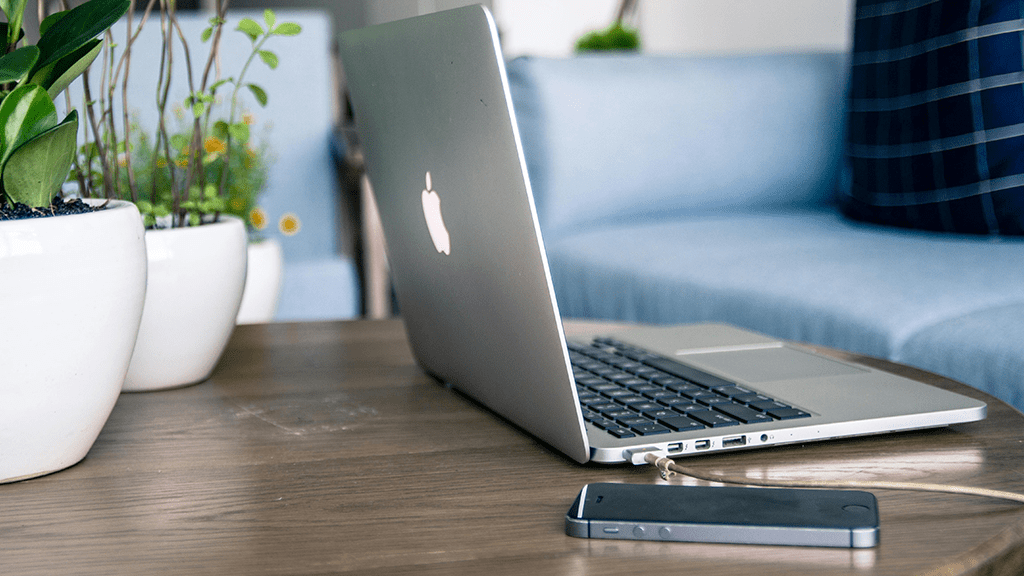 An Apple computer and smartphone sitting next to each other on a coffee table with plants and a blue couch in the background.