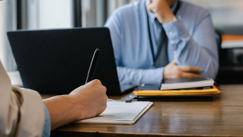 Two people seated at a desk, focused on the documents they're signing with a bold black pen.