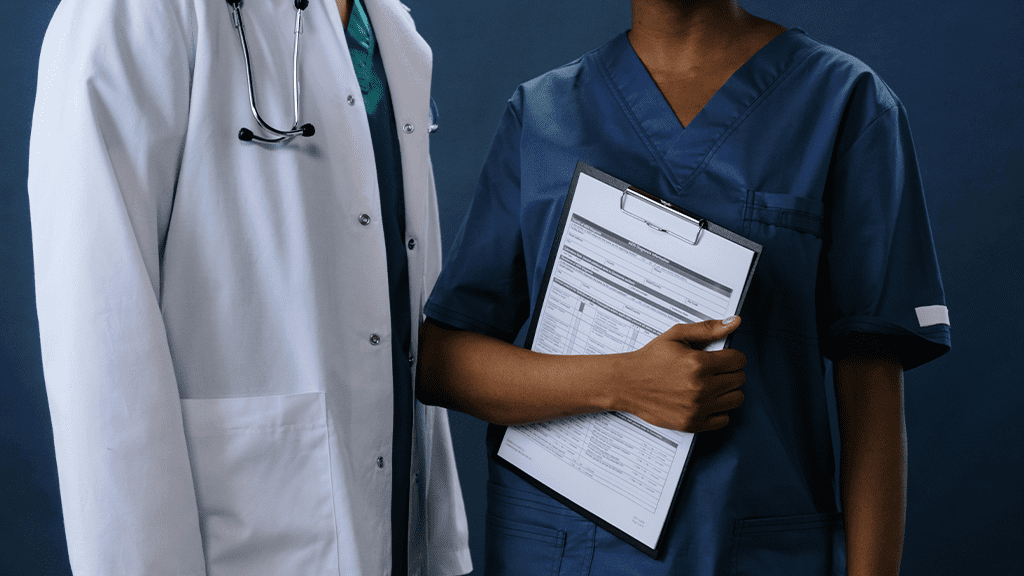 Two hospital staff, one male and one female, stand next to each other, with the female nurse holding a clip board talking about gender stereotypes.