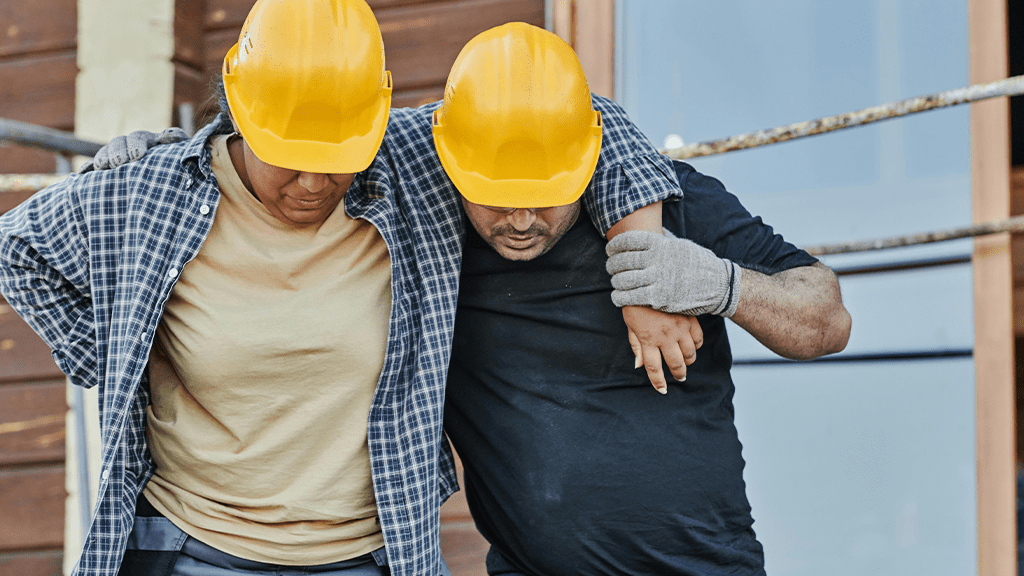 Two workers in construction hats, one worker holding onto the other as they tend to an injury off screen.