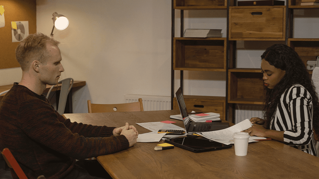 A White man and a Black woman sitting in a dimly lit room discussing an investigation and interview at a wooden table.