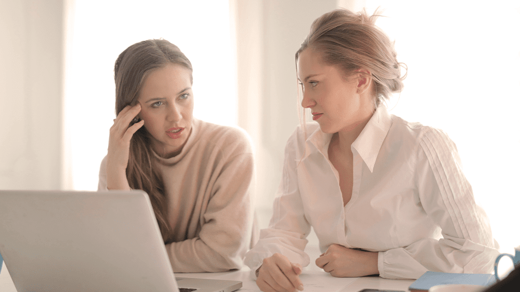 Two women seated at a working desk discussing sustainability with a laptop between them and serious thinking expressions.