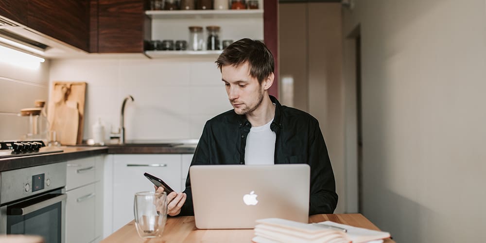Man seated at kitchen table with laptop and checking phone, searching to avoid real estate scams.