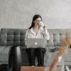 A woman seated on a modern couch sipping coffee and typing out emails on her laptop.