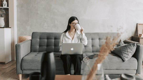 A woman seated on a modern couch sipping coffee and typing out emails on her laptop.