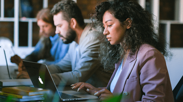 A line of people seated at a desk with their laptops open in front of them, working seriously in light of overemployment.