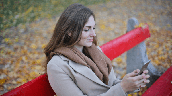 A woman sitting on a red park bench in the fall using her smartphone setting up moving.
