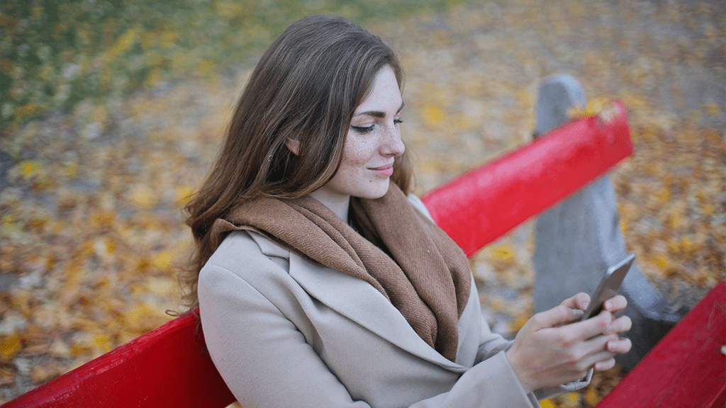 A woman sitting on a red park bench in the fall using her smartphone setting up moving.