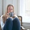 A white woman relaxes in a seat in a corner by some windows, managing stress by sipping on a cup of a nice beverage.