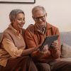 An older man and woman sit on a couch looking at a tablet together while smiling.
