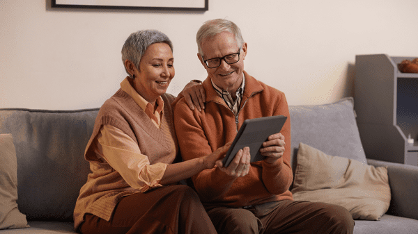 An older man and woman sit on a couch looking at a tablet together while smiling.