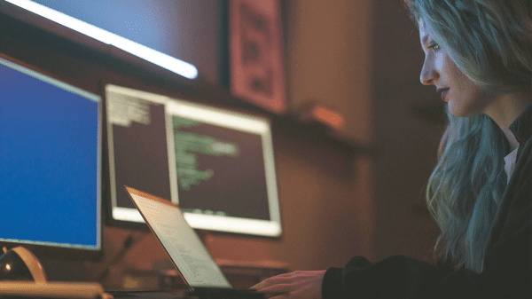 A woman with blue hair sits at a laptop with two larger desktop monitors on the desk with various code information on it to prepare for cybercrimes.