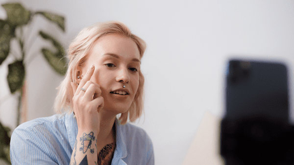 A woman with tattoos on her arm points to her face as she records in front of her phone for a Tiktok video.