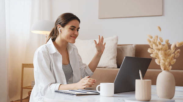 A woman waves at the laptop with airpods in as she checks in for daily stand-ups.