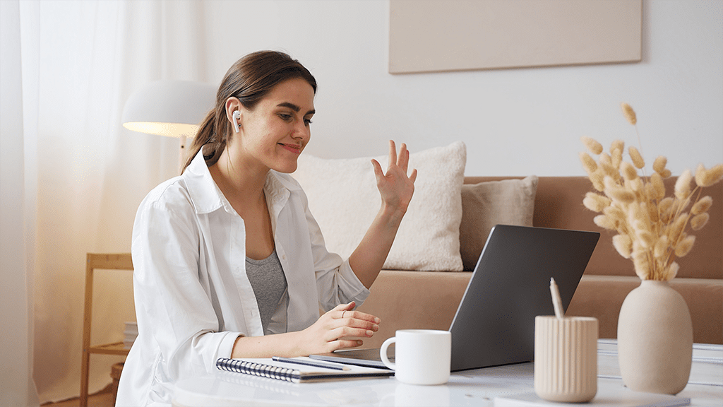 A woman waves at the laptop with airpods in as she checks in for daily stand-ups.