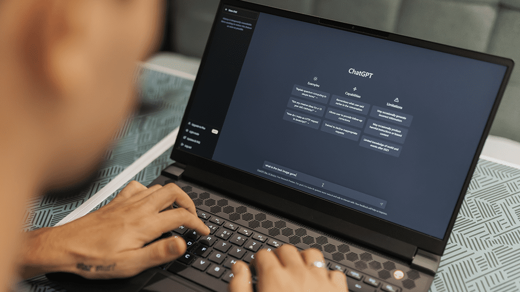 An over-the-shoulder look at a laptop open to a page of ChatGPT with a man typing into the bot's page.