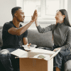 A Black man and an Asian woman high five on a couch as they unpack from their homeownership journey.