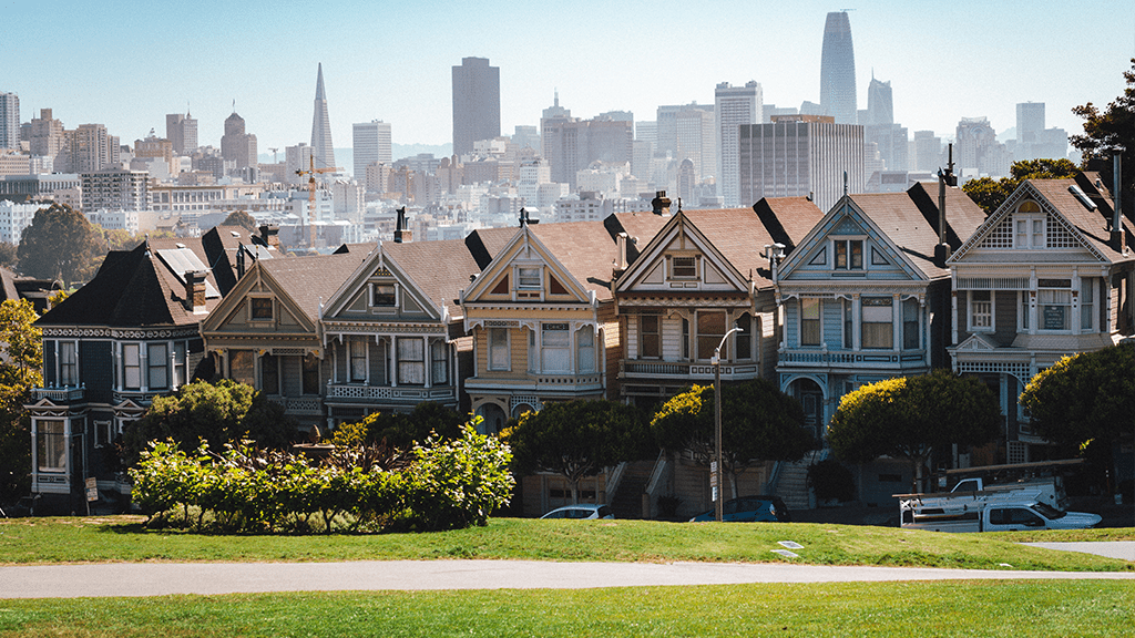 A view of houses in a suburb clustered together, with a city skyline in the distance behind the line of houses.