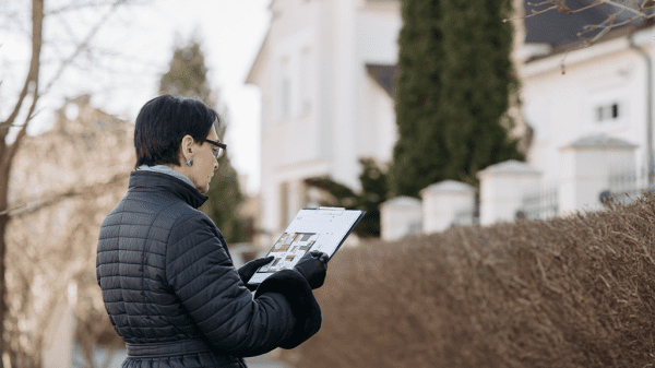 A woman stands in front of a house holding a clipboard as she reviews the documents of home sales.