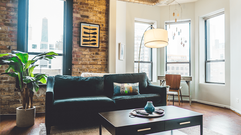 A view of an apartment with a couch, plant, and lamp, next to a window with a city view, one of many ADUs that are popping up in the LA housing market.
