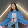 A woman stands in front of a phone on a phone stand gesturing welcomingly, with purple and yellow neon bars on the wall behind her while she records for Tiktok.