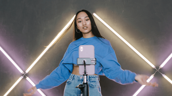 A woman stands in front of a phone on a phone stand gesturing welcomingly, with purple and yellow neon bars on the wall behind her while she records for Tiktok.