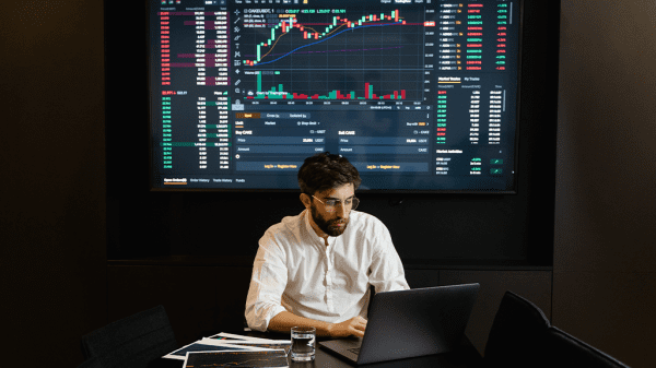 A man sits in a meeting room with a laptop and a large digital screen of charts and data on the housing market behind him.
