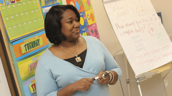 A Black female teacher stands at the front of a classroom with teaching tools around her smiling at her class.