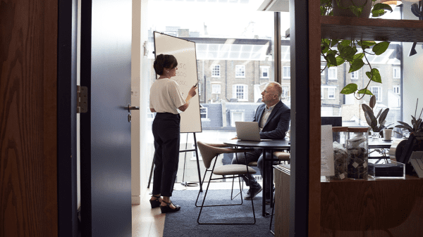 A view through an office door of a woman standing and giving a presentation to a man sitting in a desk.