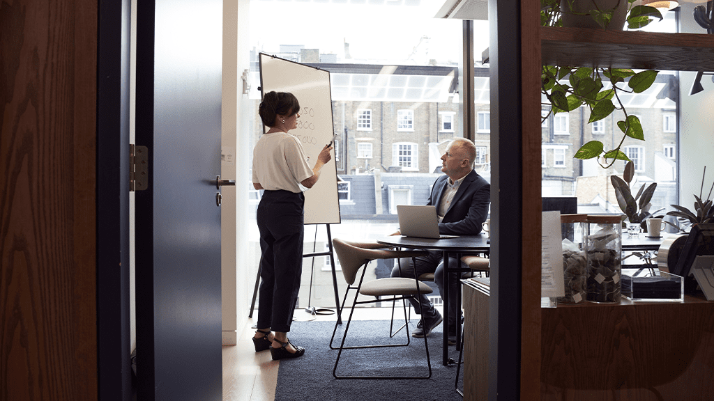 A view through an office door of a woman standing and giving a presentation to a man sitting in a desk.