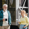 A pair of seniors, a man and a woman, standing outside of a home with mugs of a drink in their hand, laughing as they talk.